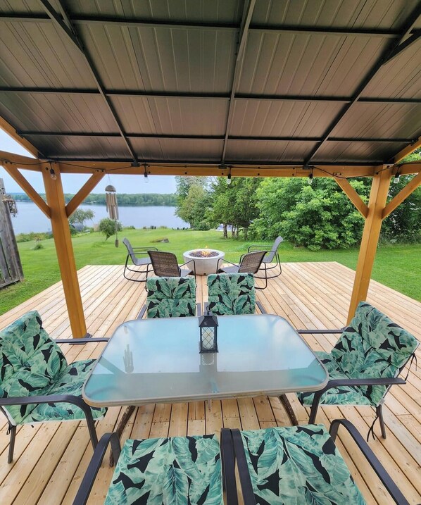 Una terraza exterior con una mesa y sillas de vidrio, rodeada por una pérgola de madera, con vista a un lago.