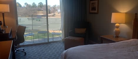 Ett hotellrum med en säng, ett skrivbord, en stol, ett litet bord och utsikt över en tennisbana genom en glasdörr.