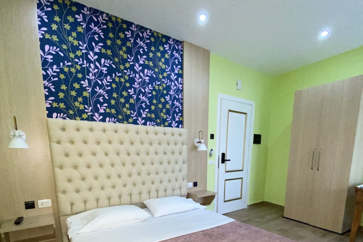 Un dormitorio con una cama grande, un cabecero, una mesita de noche, un armario y una pared con estampado floral.
