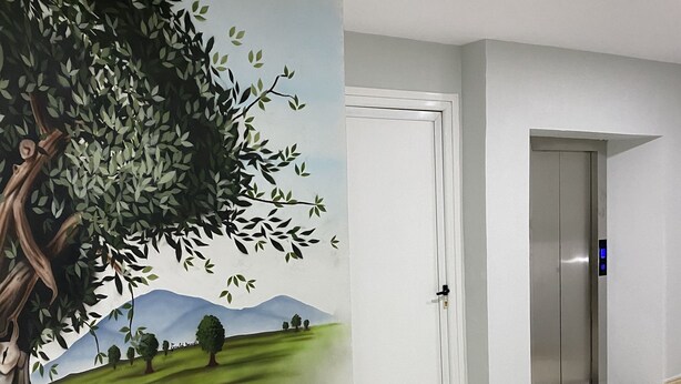 Un couloir orné d’une fresque représentant un arbre et un paysage, avec une porte blanche et un ascenseur moderne.
