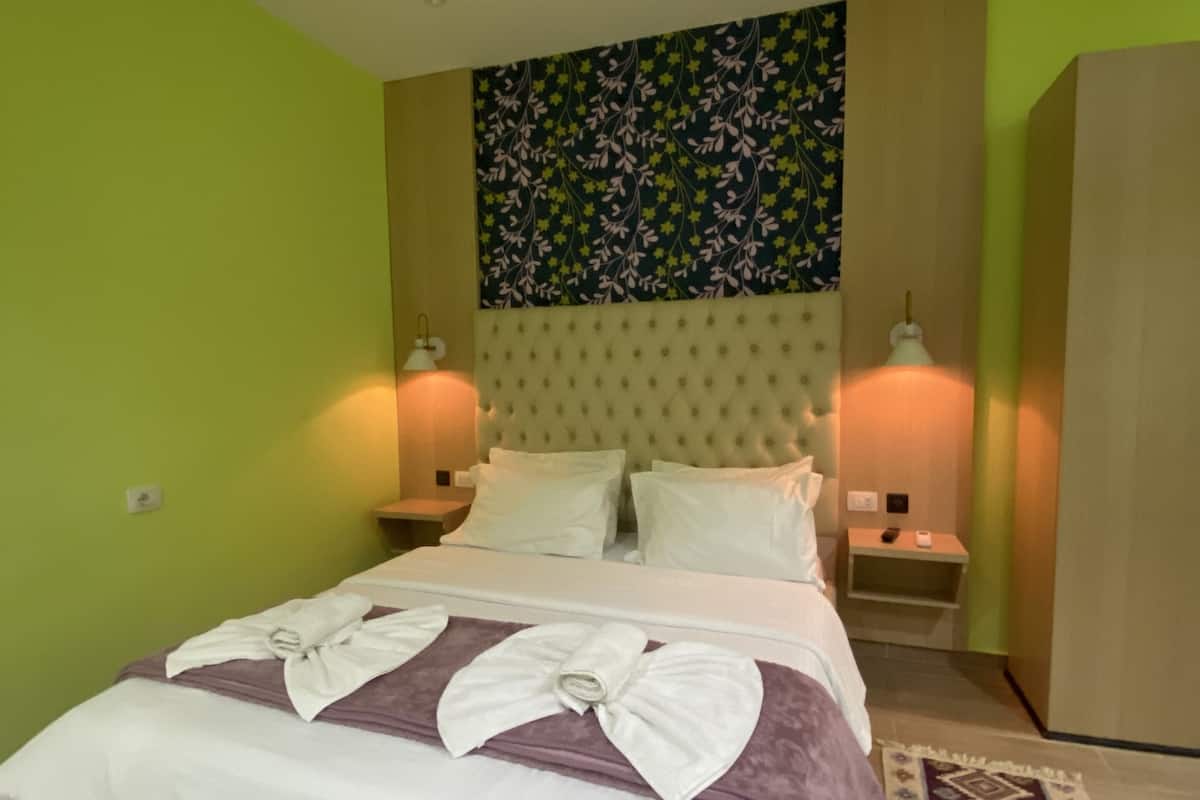 Una cama bien hecha con sábanas blancas y una manta morada, dos lámparas de noche, un cabecero estampado con motivos florales y una pared verde.