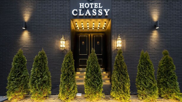 Entrada de un hotel con fachada de ladrillo oscuro, un letrero que dice "HOTEL CLASSY" y una fila de árboles de Navidad al frente.