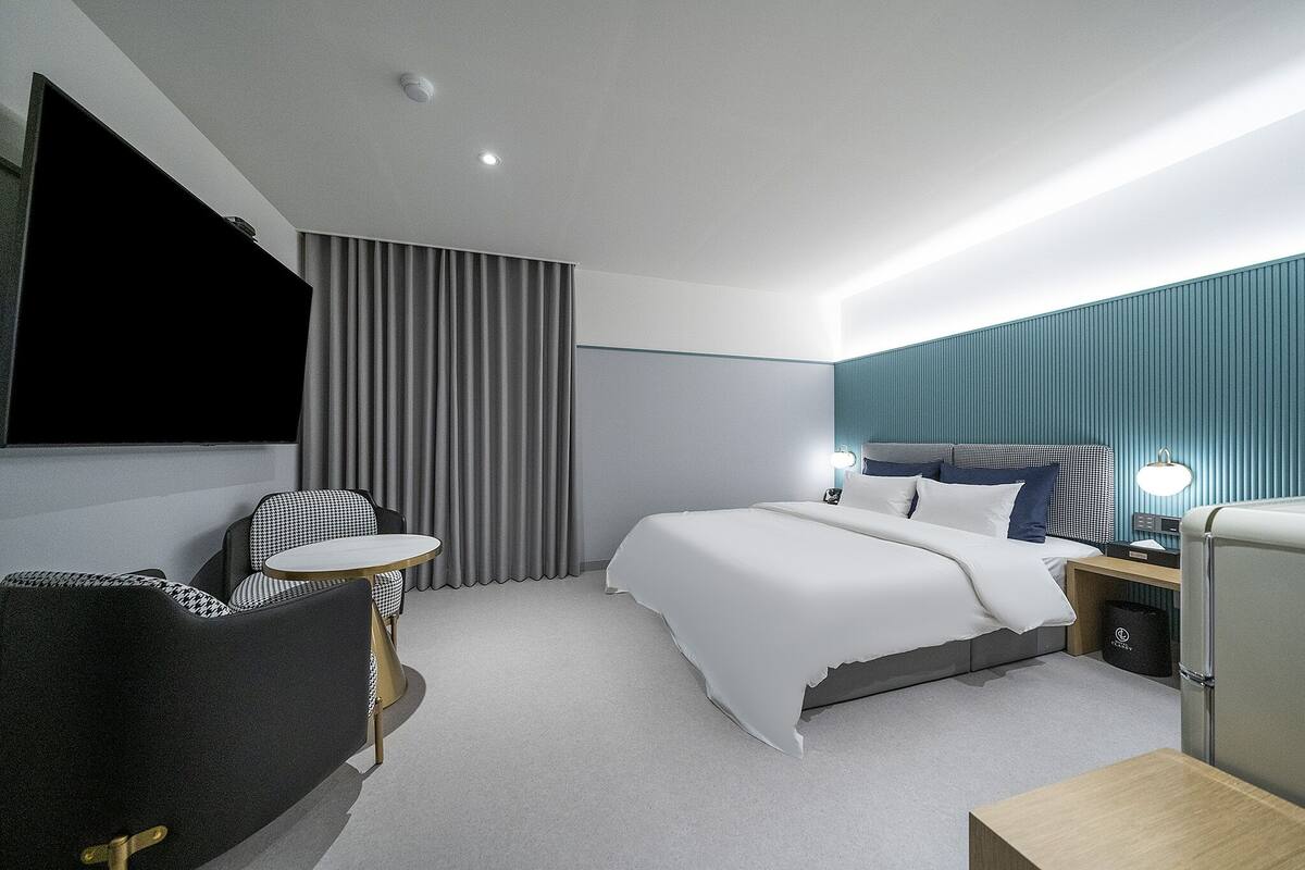 A modern hotel room with a bed, a small round table, a chair, a TV, and a nightstand with a lamp.