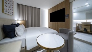 Una habitación de hotel moderna con una cama grande, una mesita de noche, un televisor y un baño visible a través de una puerta abierta.