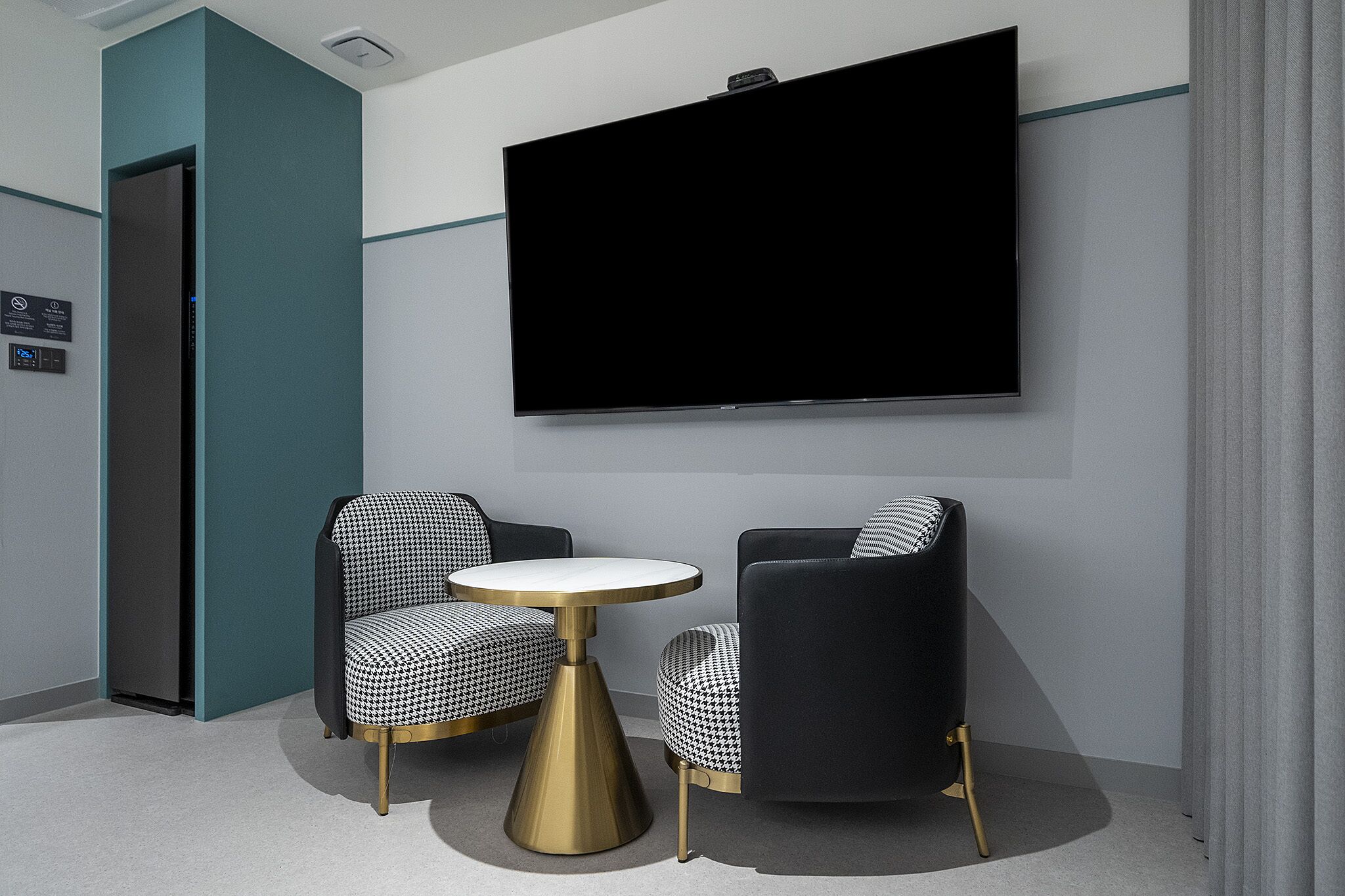 Une chambre moderne dotée d’un téléviseur à écran plat fixé au mur, de deux fauteuils à motifs et d’une petite table ronde avec un pied doré.