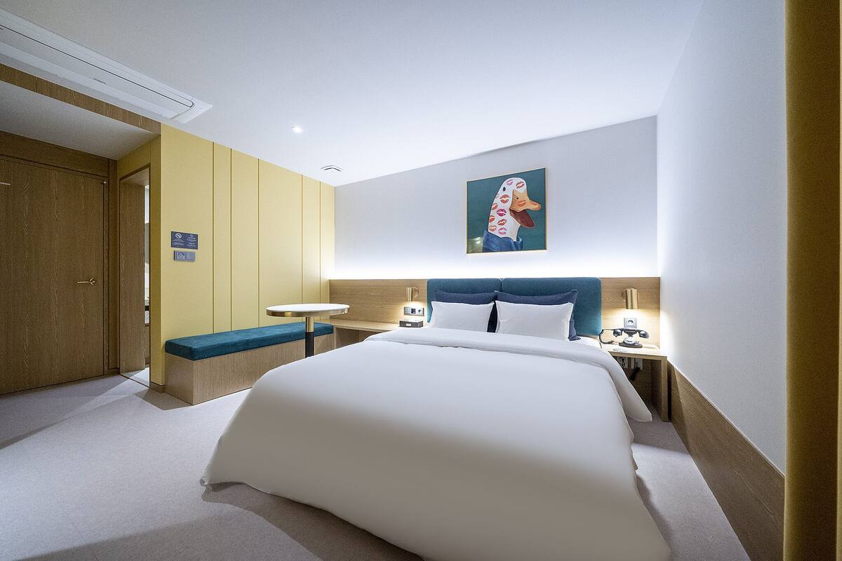 A hotel room with a large bed, a bedside table with a lamp, a small round table, and a bench.