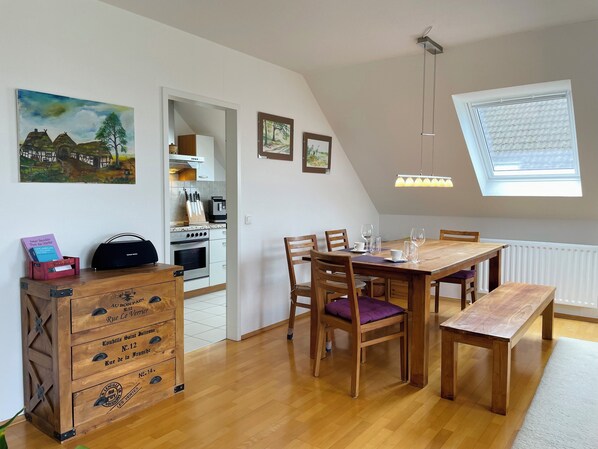 Un comedor con una mesa de madera, sillas y un banco. Se observa una cocina al fondo con estufa y armarios.
