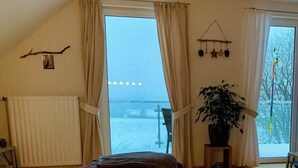 A cozy living room with a large window, a purple ottoman, a wooden table with a dish, and a potted plant. - FeWo Siebenstern Fernblick am Hohen Venn (Monschau)