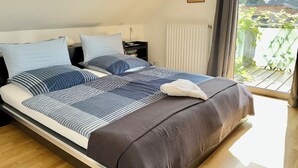 A modern bedroom with a large bed, a nightstand, a window with a view of greenery, and a ceiling with a sloped design. - FeWo Siebenstern Südblick am Hohen Venn (Monschau)