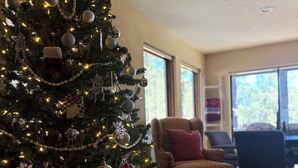Un árbol de Navidad con adornos y luces, un sillón estampado, y una mesa de comedor en una habitación con suelo de madera y ventanas amplias.
