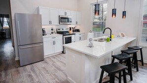 Una cocina moderna con armarios blancos, electrodomésticos de acero inoxidable, una isla central con taburetes e iluminación colgante.
