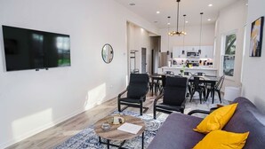 Un salón moderno con televisor de pantalla plana, un sofá con cojines amarillos, una mesa de centro y una zona de comedor con sillas, además de una cocina al fondo.