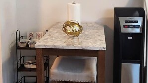 Un comedor con una mesa de mármol, un adorno dorado, una silla, un reloj de pared, un dispensador de agua y una estantería metálica.