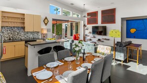 Een moderne keuken met houten kastjes, een eethoek met een ronde tafel, grijze stoelen, en een groot kookeiland met een spoelbak.