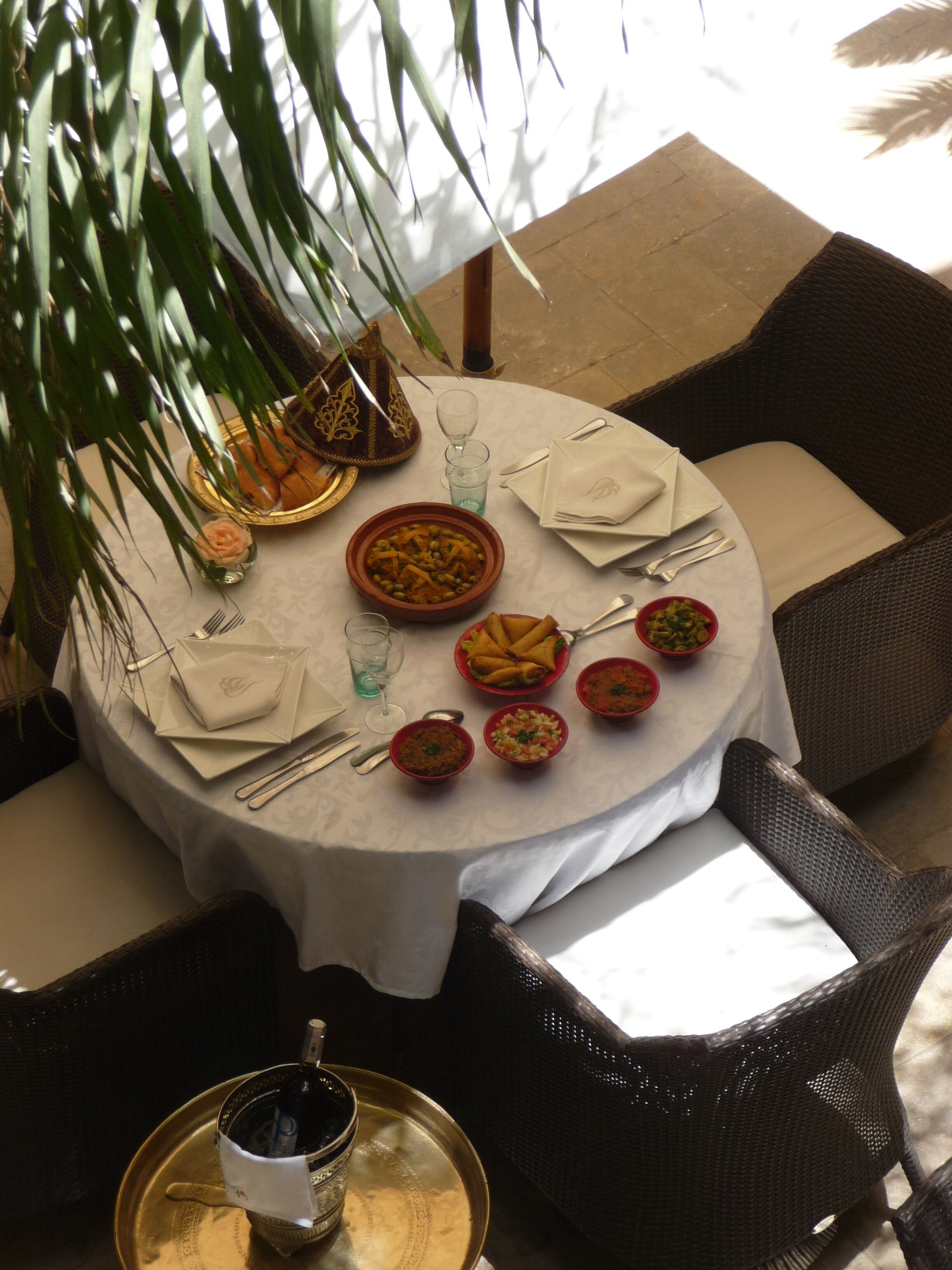 A round dining table with a white tablecloth, surrounded by wicker chairs. There are plates of food, a glass of water, and a bottle of water on the table.