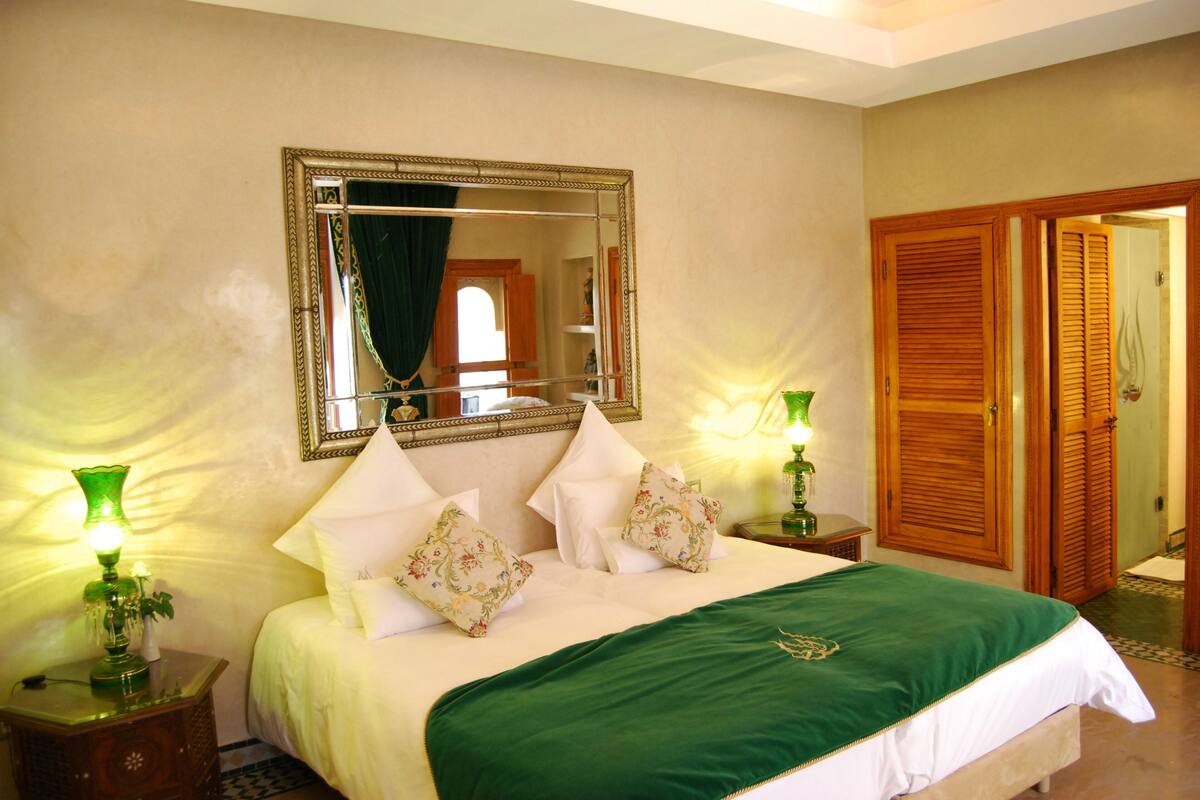 Cama bem arrumada com colcha verde, duas luminárias de apoio e um espelho grande em cima.