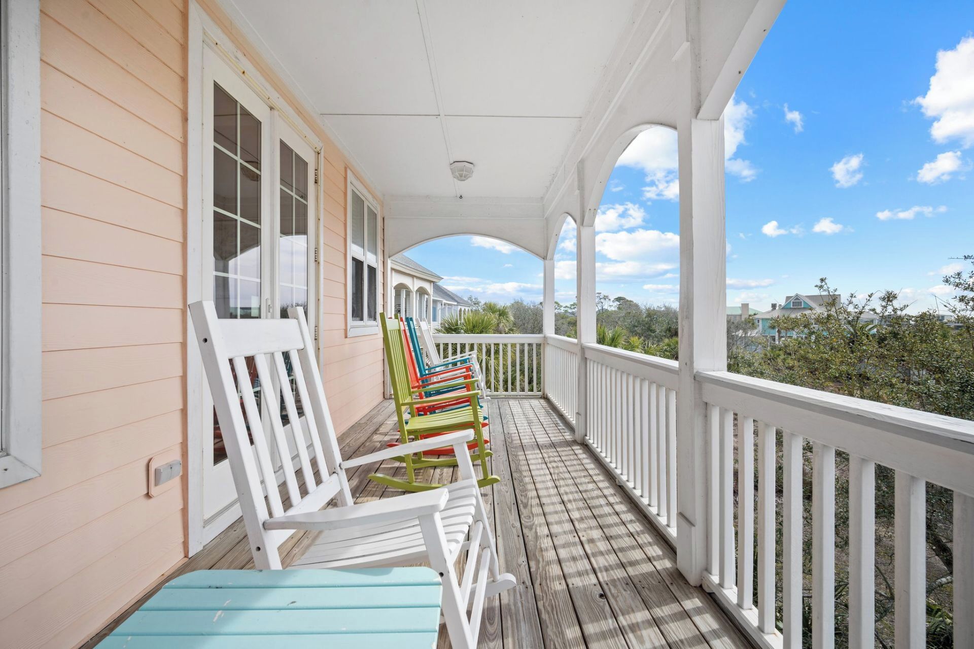 A covered porch with white railings and a wooden deck, featuring several colorful Adirondack chairs.