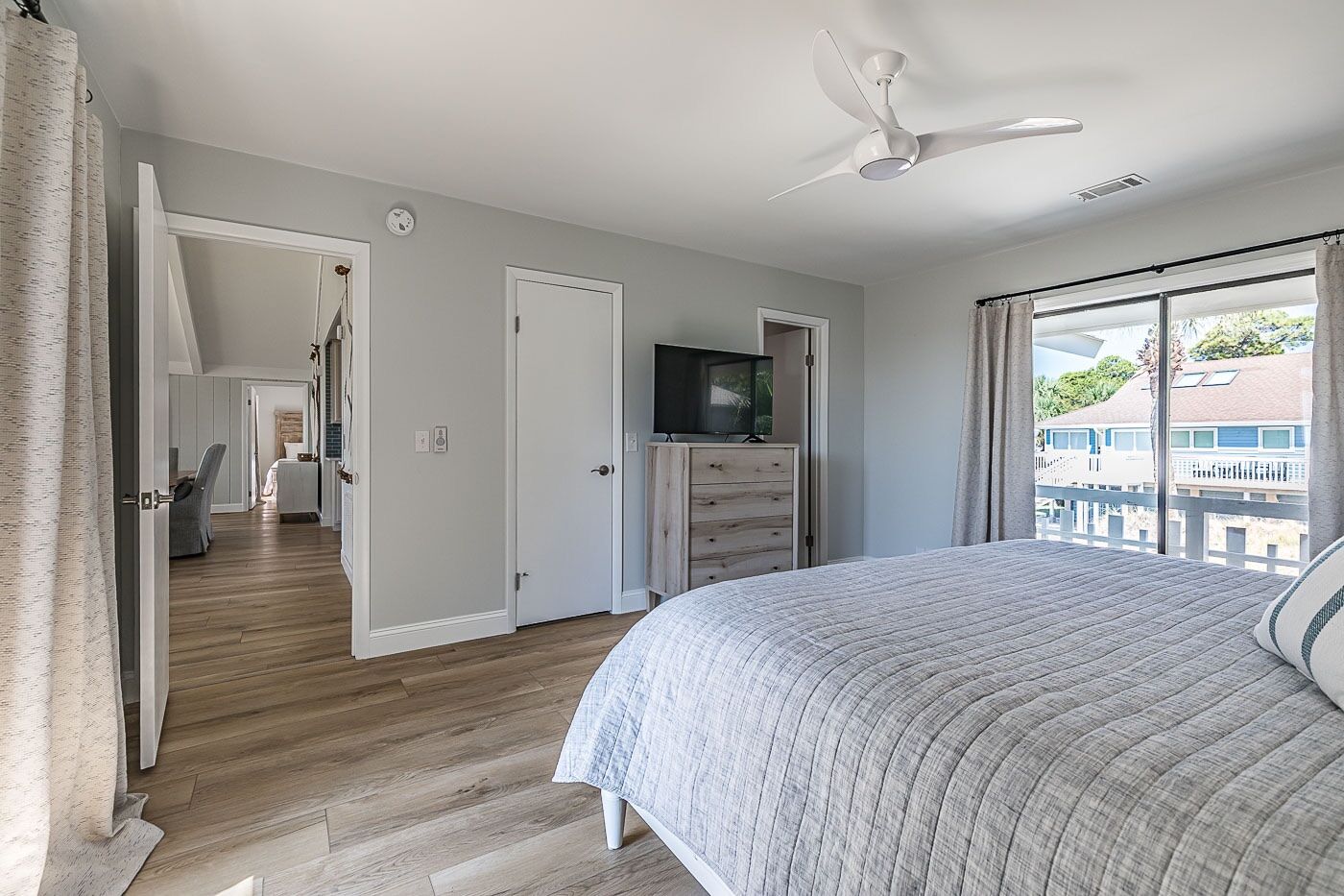 A bedroom with a large bed, a ceiling fan, a TV, and a view of the outside.