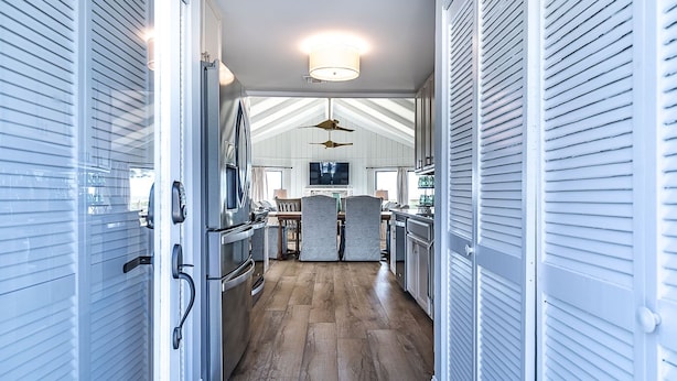 A narrow hallway with a kitchen area at the end, featuring a large refrigerator, wooden flooring, and a ceiling fan.