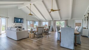 A spacious living room with a high ceiling, wooden flooring, a television, and a dining area with chairs and a table.