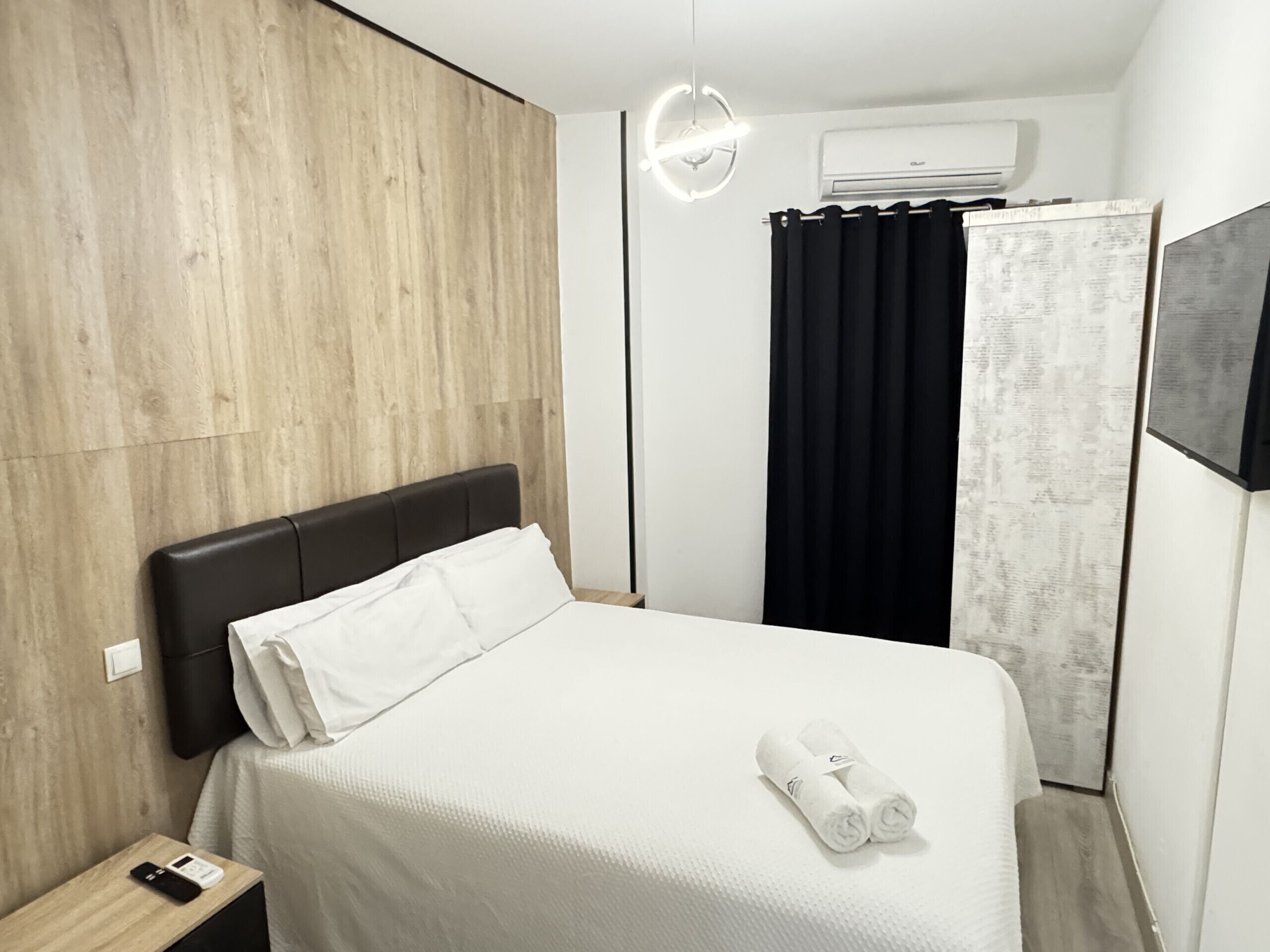 A hotel room with a wooden panel wall, a bed with white bedding, a black headboard, a bedside table with a phone, and a wall-mounted air conditioner.