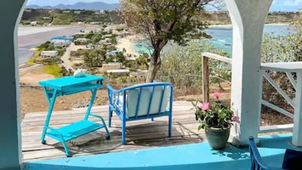 A covered outdoor area with a view of a beach and buildings. There is a blue table and chair set, and a potted plant. - Villa Kaiae (The Valley)