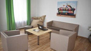 Een moderne woonkamer met een beige bank, twee fauteuils, een houten salontafel met een laptop, een groen gordijn en een kleurrijk schilderij aan de muur.