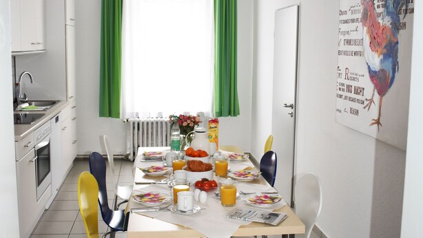 Een eetruimte met een tafel voor vier personen, stoelen, een raam met groene gordijnen en een keuken met witte kasten.