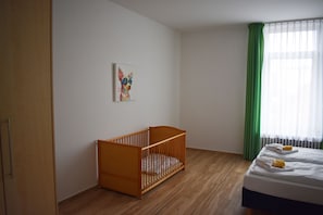 Une pièce avec un berceau en bois, un lit avec des serviettes, un tableau au mur et une fenêtre avec des rideaux verts.