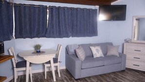 Una habitación con un sofá gris, una mesa blanca con sillas, un televisor montado en la pared y techo de madera.