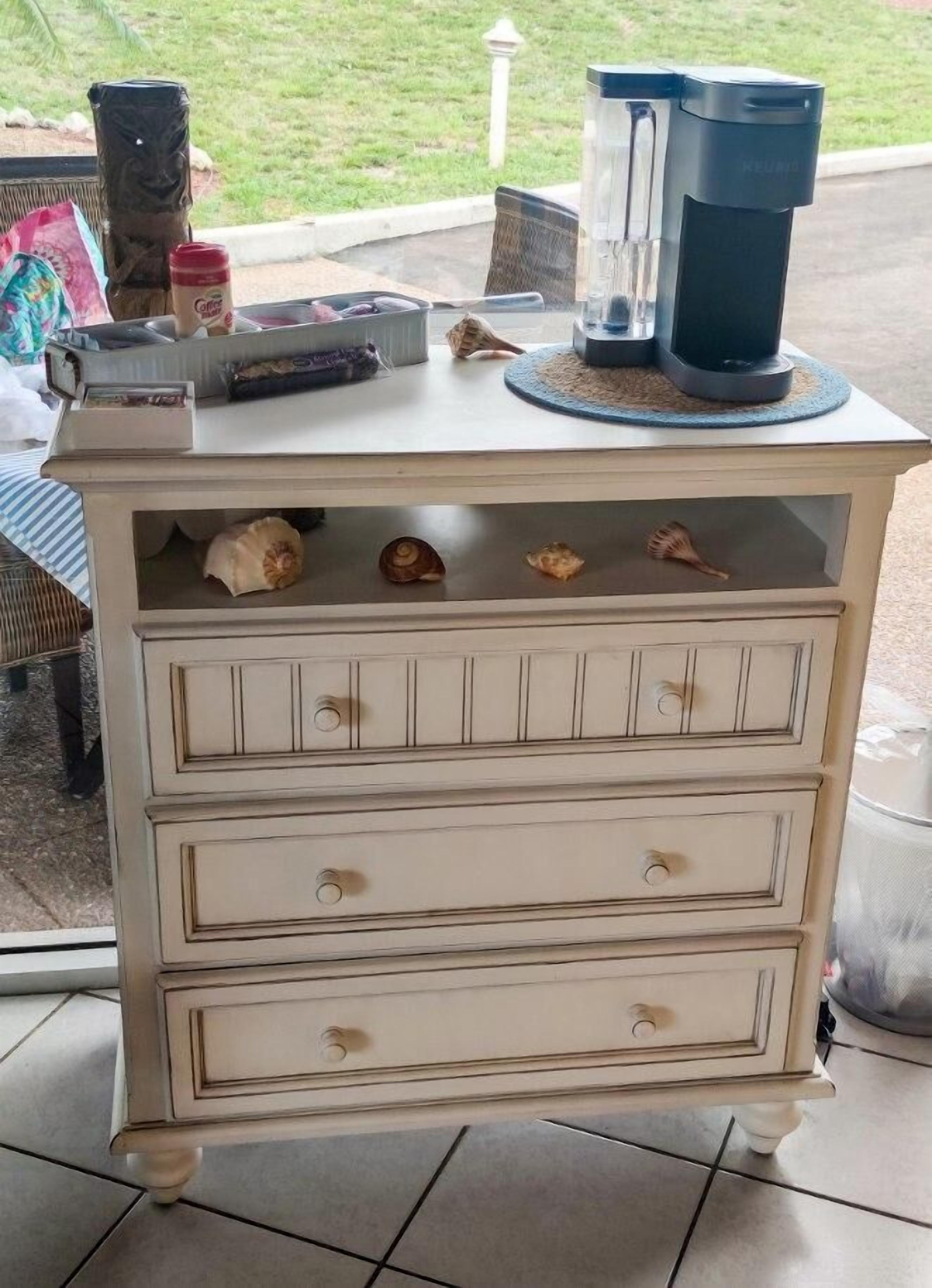 Une commode blanche avec des tiroirs, une machine à café et des coquillages décoratifs posés sur le dessus.