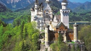 Um castelo com várias torres, rodeado por vegetação exuberante e montanhas.