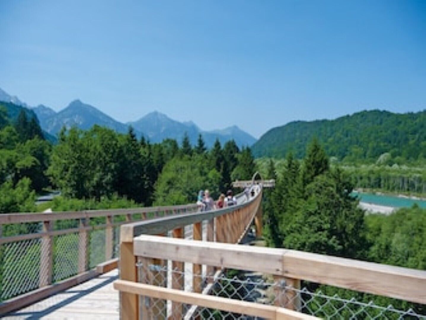 A wooden suspension bridge with a chain-link fence, overlooking a mountainous landscape with dense forests and a clear blue sky.