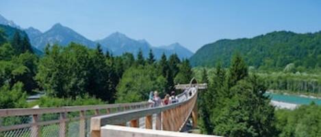 A wooden suspension bridge with a chain-link fence, overlooking a mountainous landscape with dense forests and a clear blue sky.