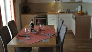 Uma sala de jantar com mesa de madeira posta para dois, uma garrafa de vinho e copos. Cadeiras cercam a mesa, e uma área de cozinha é visível ao fundo.