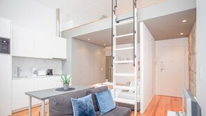 A modern apartment with a kitchenette, a grey sofa with blue cushions, a small dining table, and a loft-style sleeping area with a ladder.