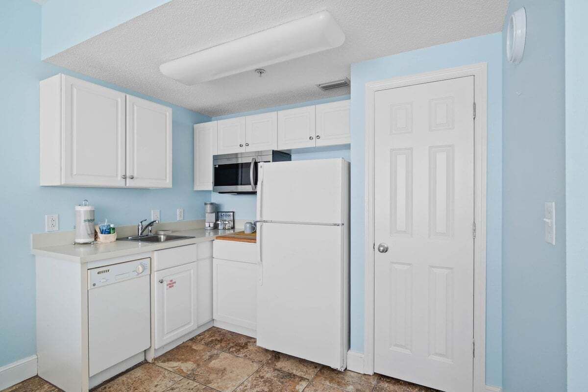 Una cocina compacta con armarios blancos, microondas, refrigerador y lavavajillas.