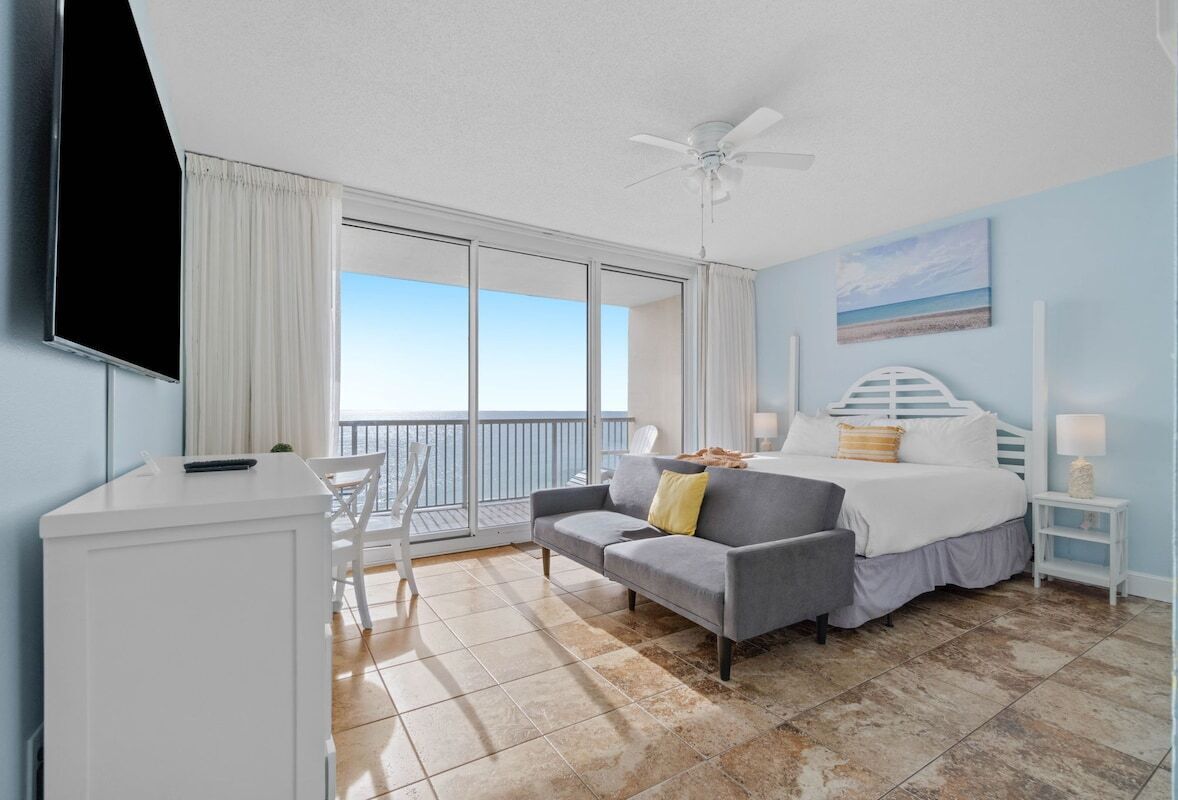 Habitación de hotel con cama, escritorio, silla, ventilador de techo y un ventanal con vista al mar.