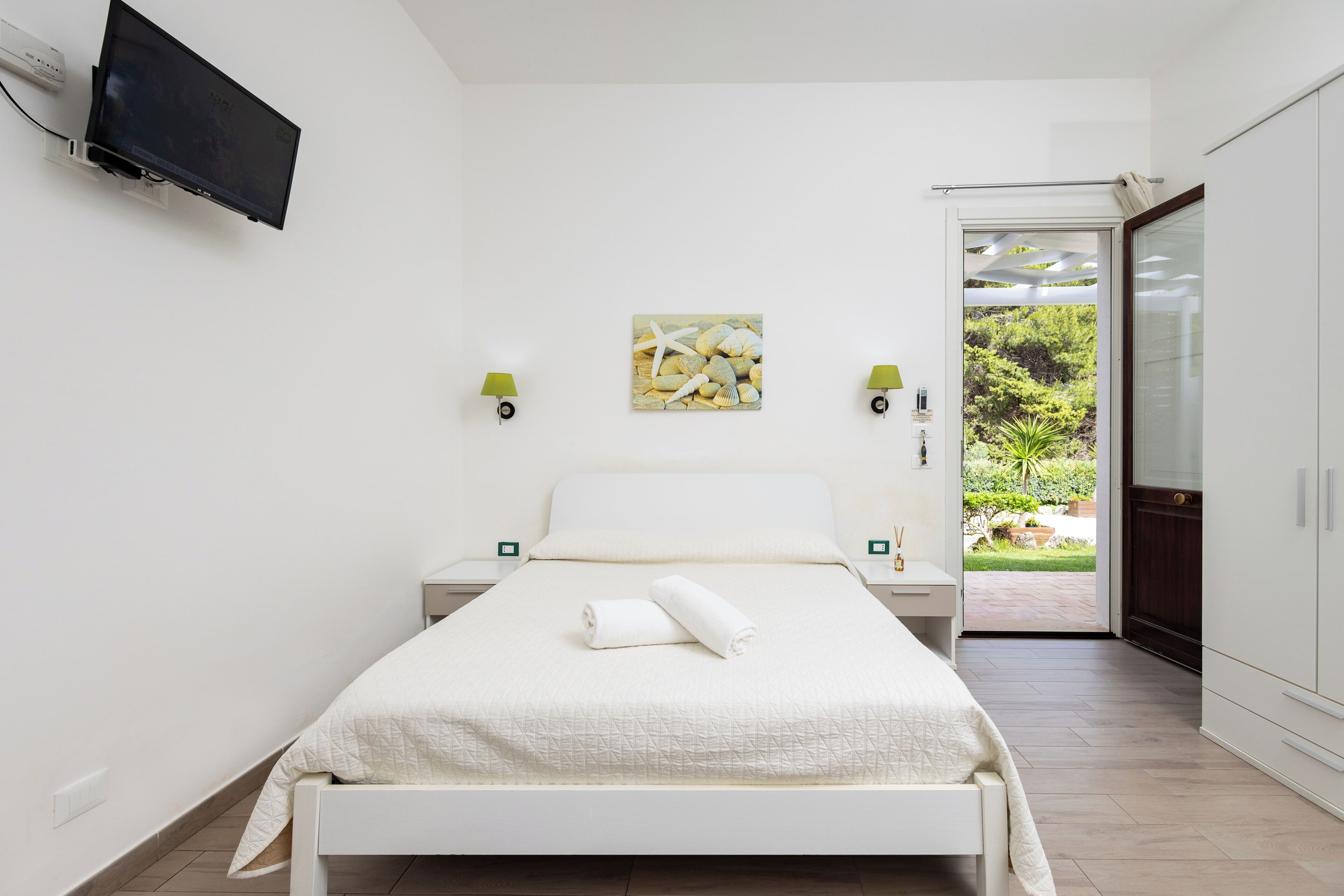 A bedroom with a bed, a TV mounted on the wall, a painting, and a view of the outdoors.