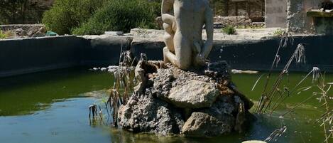 Una estatua de piedra de una figura sentada sobre una roca, en medio del agua, rodeada de vegetación y montañas al fondo.