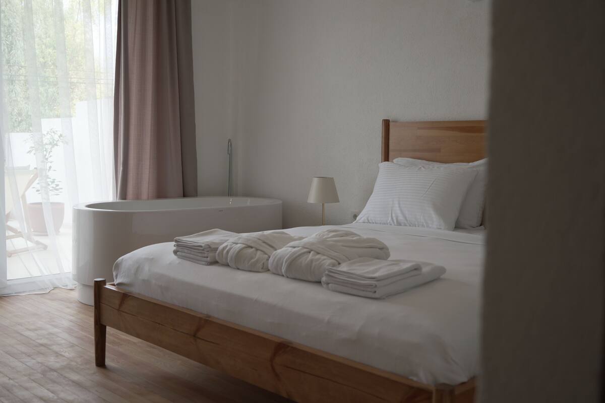 A neatly made bed with folded towels, a bedside table with a lamp, and a view of the outdoors through sheer curtains.