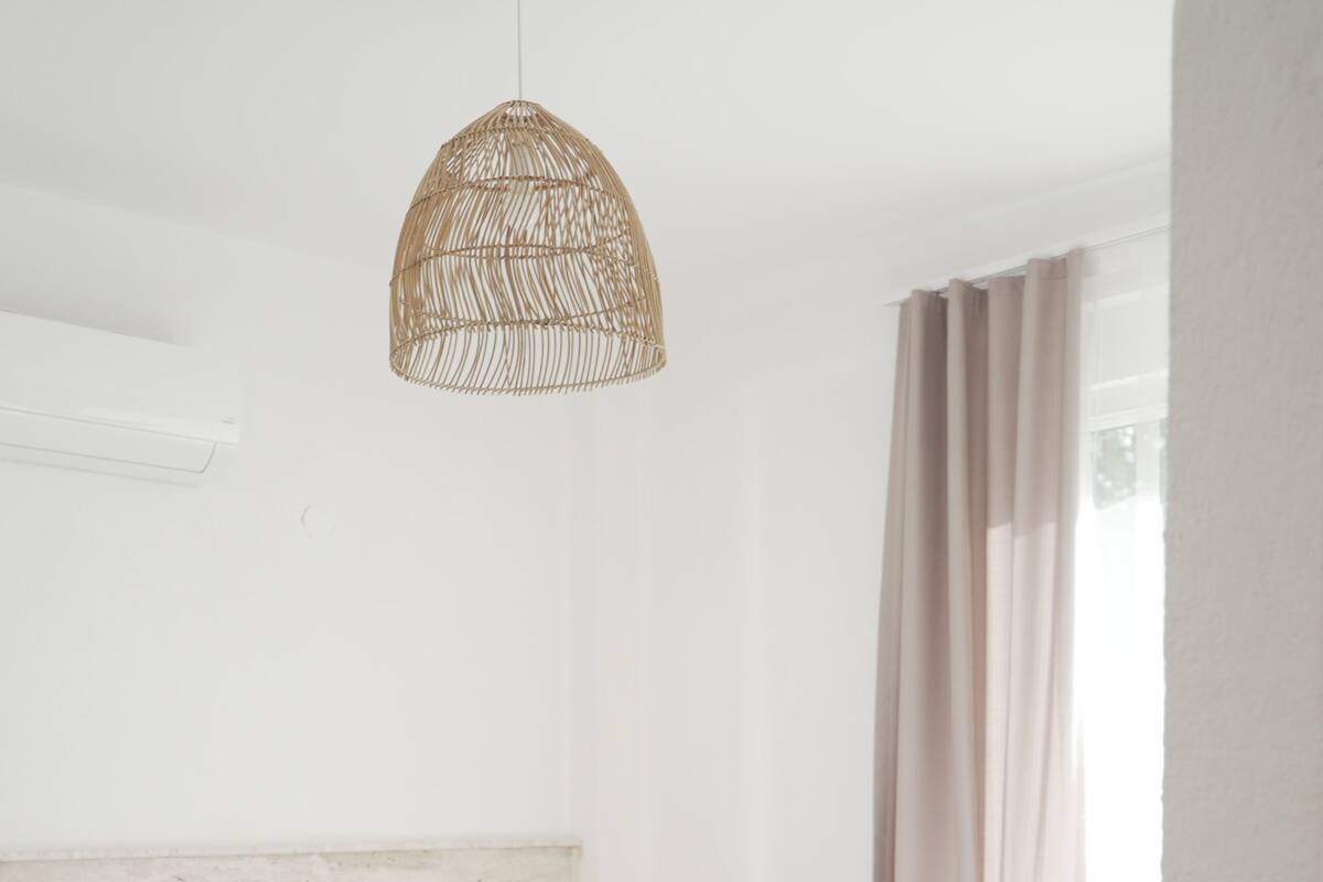 A modern bedroom with a wooden bed, white linens, a woven pendant light, and a white bathtub.