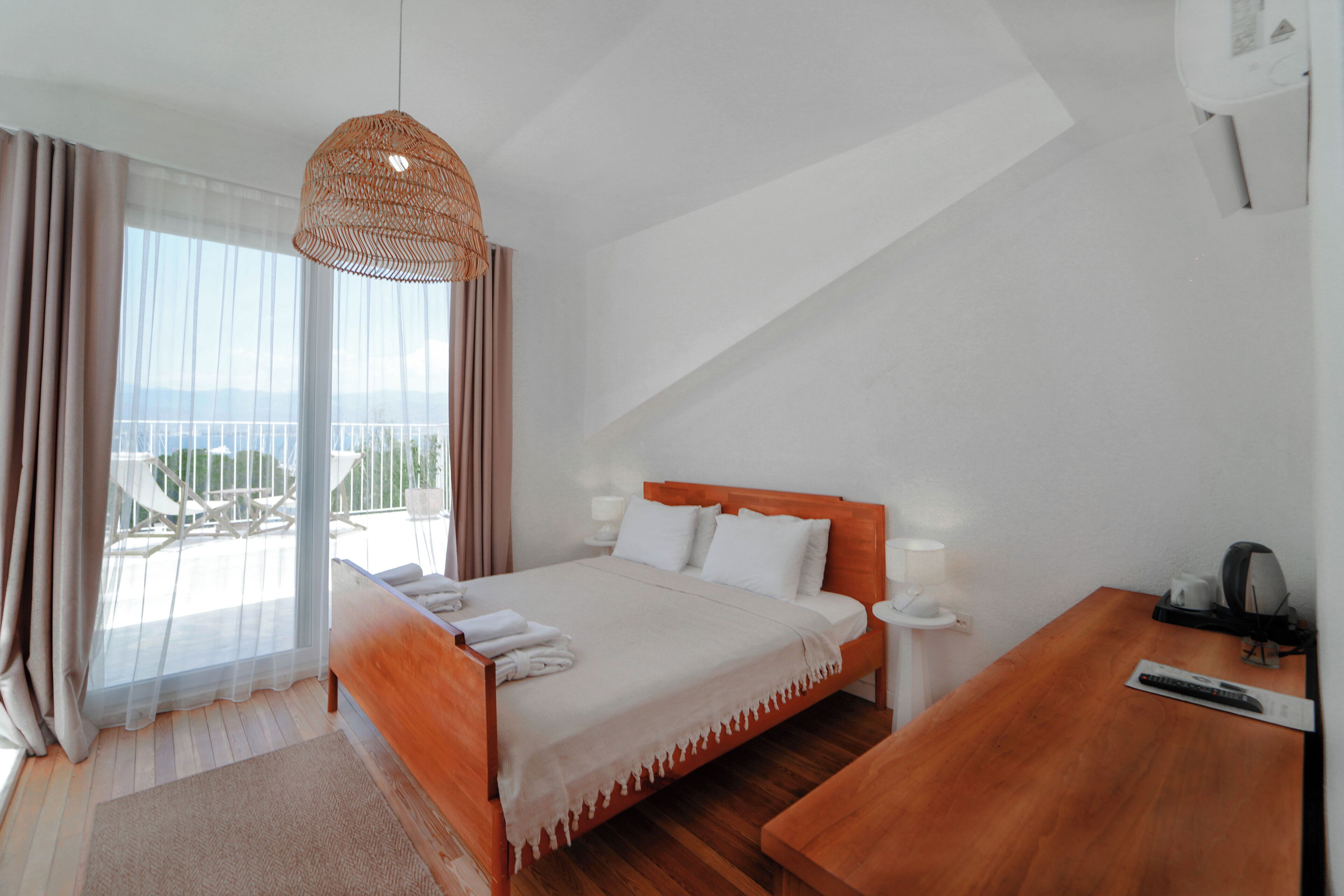 A bedroom with a bed, bedside tables, a wooden desk, a hanging light fixture, and a view of the outdoors.