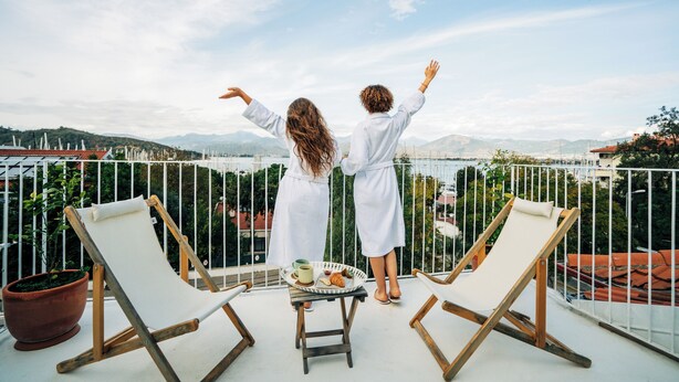 Un balcón con dos sillones de descanso, una mesa con un croissant y una taza, y vistas a montañas y edificios.