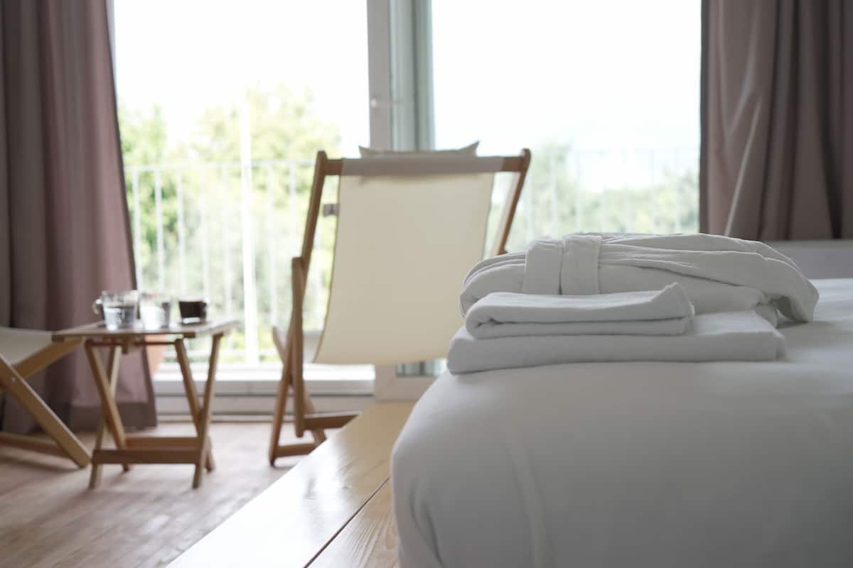 A hotel room with a neatly made bed, a small table with a chair, and a view of greenery outside.