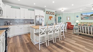A spacious kitchen with white cabinets, a wooden island, and a dining area with white chairs. - Just Coasting - Saltwater Pool, 5 Bedrooms, Across Street to Beach (North Myrtle Beach)
