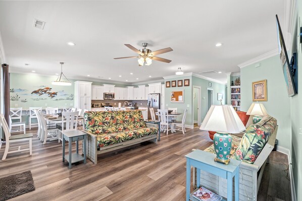 A spacious living area with a sofa, dining table, and chairs, a kitchen in the background, and a ceiling fan. - Just Coasting - Saltwater Pool, 5 Bedrooms, Across Street to Beach (North Myrtle Beach)