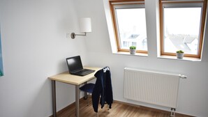 Une pièce comprenant un bureau en bois, un ordinateur portable, une chaise, un radiateur et deux fenêtres ornées de plantes en pot.