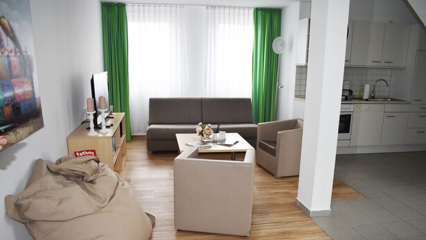 Un salon moderne avec un canapé, une table basse et un fauteuil en forme de poire. On aperçoit un coin cuisine à l’arrière-plan.