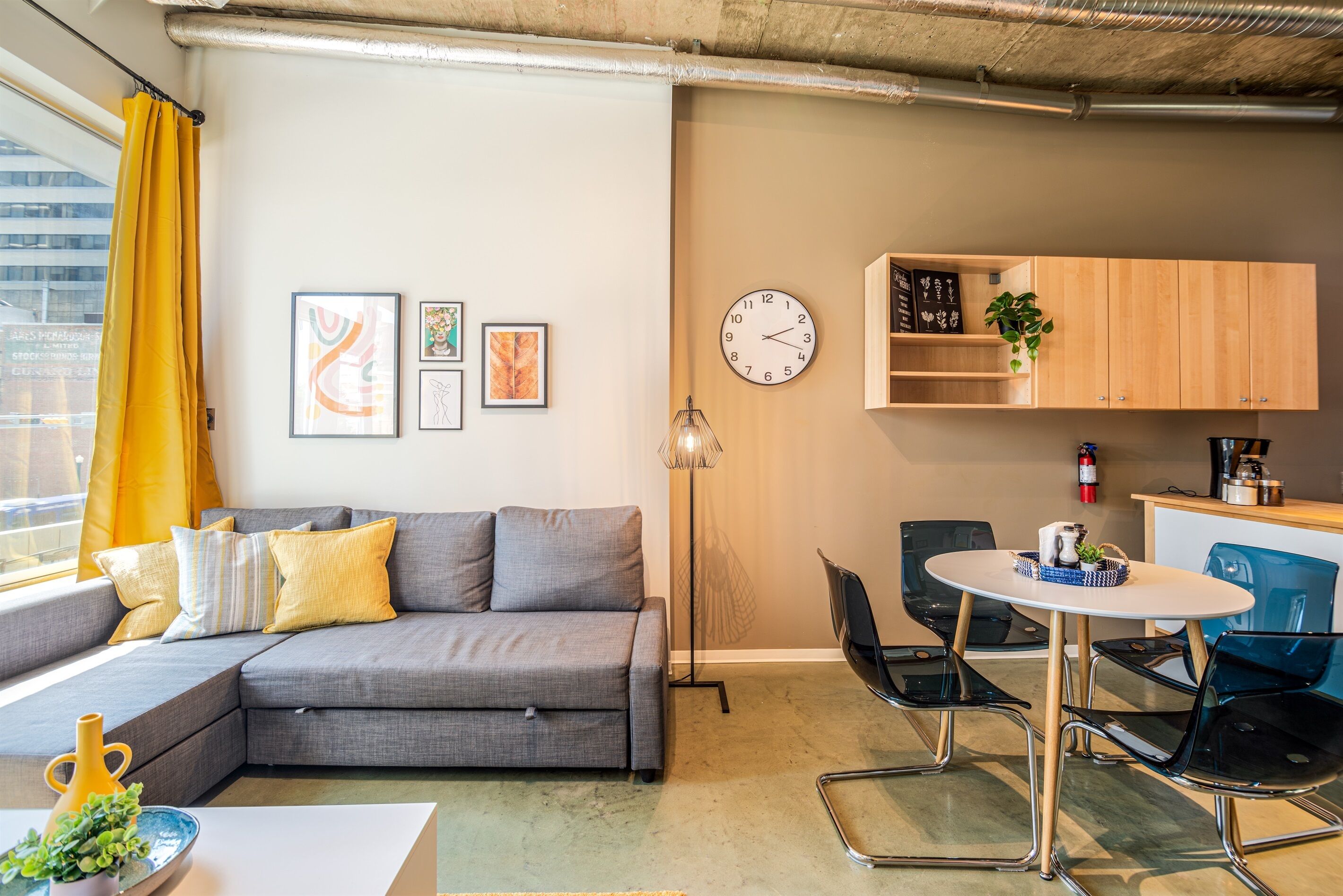 Una sala de estar moderna con un sofá gris, cojines amarillos, una mesa redonda con sillas, una lámpara de pie y un reloj en la pared.